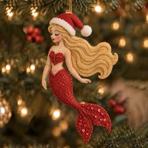May include: A felt mermaid ornament featuring long blonde hair, a red Santa hat with a white pom-pom, a red heart-shaped top, and a shimmering red sequined tail. The figure is suspended by a gold string against a blurred Christmas tree.