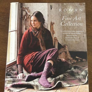 May include: A book cover titled "Rowan Fine Art Collection" featuring a woman wearing a burgundy outfit and socks. The cover text indicates 14 sock and accessory designs for women. The background shows a rustic interior with a window.