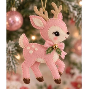 May include: A felt deer ornament in shades of light pink and cream with gold glitter antlers. The deer features a pink bow with a small gold bell and green felt holly leaves at the neck, white spots on the back, and dark brown hooves.