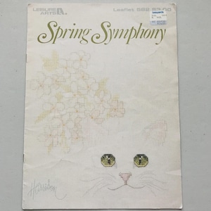 May include: A Leisure Arts cross-stitch pattern leaflet titled Spring Symphony, Leaflet 582. The cover features a white cat with green eyes and a pink nose, partially obscured by a cluster of yellow and white flowers, with the artist name Harrison.