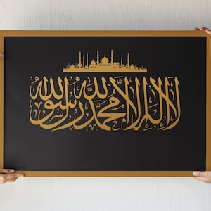 May include: A framed art print featuring gold Arabic calligraphy and a mosque silhouette on a black background. The frame is a warm gold color. The artwork is held by two pairs of hands.
