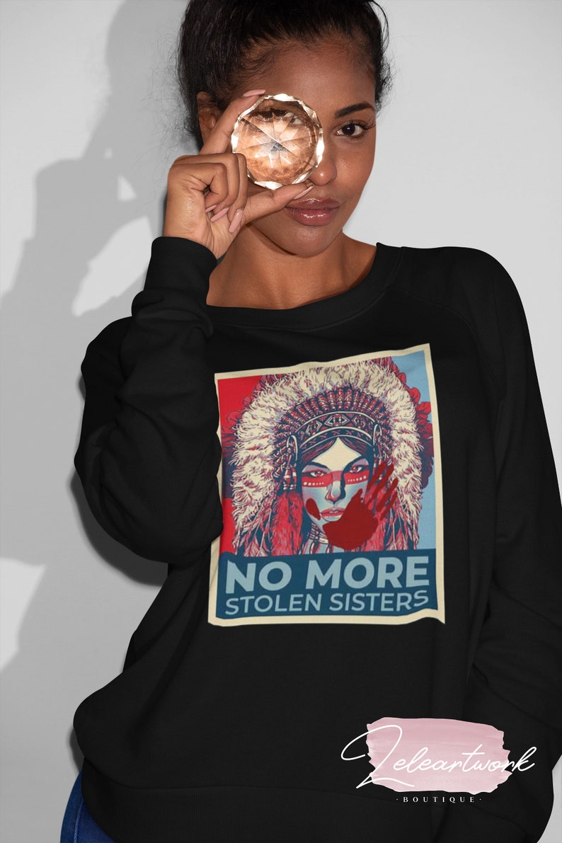 May include: Black long-sleeved sweatshirt with a graphic of a Native American woman in a headdress and the text "NO MORE STOLEN SISTERS." The design uses red and blue colours. The sweatshirt has a crew neck.