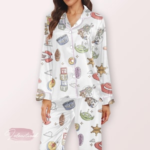 May include: White pajama set featuring a colorful Toy Story print. The long-sleeved top and pants are adorned with cartoon characters, stars, and other themed graphics. The set has pink trim.