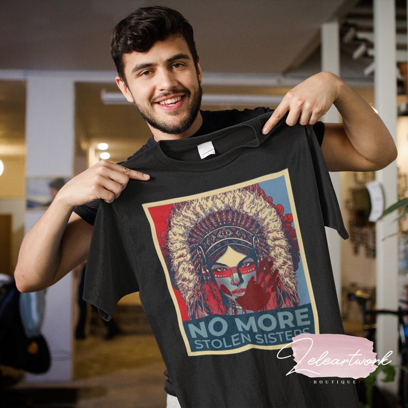 May include: Black t-shirt with a graphic of a Native American woman in a headdress, and the text "NO MORE STOLEN SISTERS". The design incorporates red, blue, and beige colours.