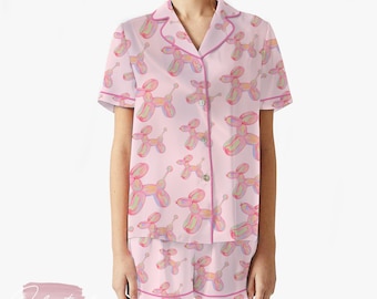 May include: A light pink pajama set with a repeating pattern of balloon dogs in pink, yellow, and green. The set includes a short-sleeved button-down shirt and matching shorts, both trimmed with pink piping.