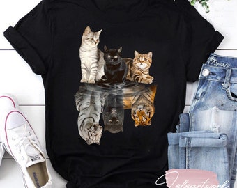 Può includere: Una maglietta nera con una grafica di quattro gatti riflessi nell'acqua come tigri. I gatti sono un tabby, un gatto nero e due tabby arancioni. Le tigri sono riflesse capovolte.