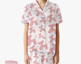 May include: White pajama set with a pink balloon dog pattern. The set includes a short-sleeved button-down shirt and matching shorts. The balloon dog design features pink, yellow, and purple accents. The pajama set is made of a soft, lightweight fabric.