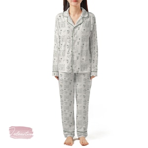 May include: Light grey pyjama set featuring a repeating pattern of castles, flowers, clouds, and envelopes. The long-sleeved top and trousers have dark green trim. The pyjamas are made of a soft, lightweight fabric.
