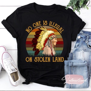 Peut inclure: T-shirt noir avec un graphique vintage d'un Am&eacute;rindien portant une coiffe. Le graphique est sur un fond de coucher de soleil avec le texte "No One Is Illegal On Stolen Land".