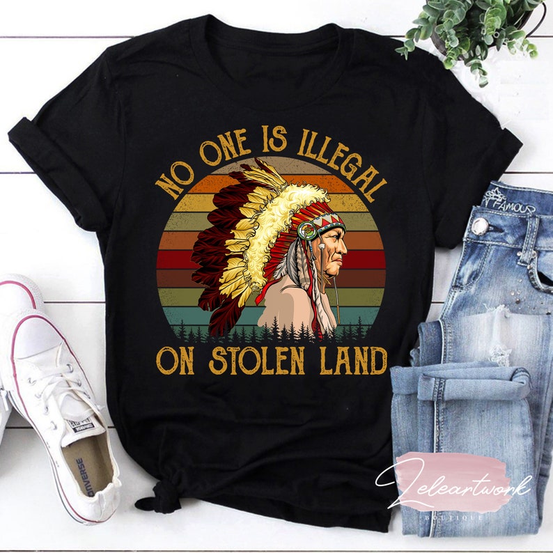 Peut inclure: T-shirt noir avec un graphique vintage d'un Am&eacute;rindien portant une coiffe. Le graphique est sur un fond de coucher de soleil avec le texte "No One Is Illegal On Stolen Land".