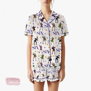 Könnte beinhalten: Weißes Pyjama-Set mit einem sich wiederholenden Muster aus stilisierten Figuren, Kronen und dem Wort "SIX" in Lila. Das Set beinhaltet ein kurzärmeliges Oberteil und passende Shorts, die mit lila Paspeln versehen sind.