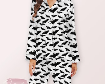 May include: White pajama set with a black bat pattern. The long-sleeved top has a button-down front and a notched collar. The pants are long and have a matching bat print. The pajamas are made of a soft, silky material.