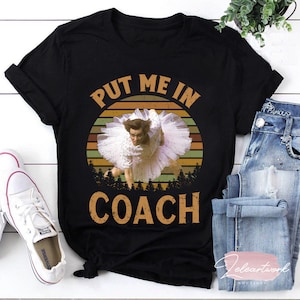 May include: Black t-shirt featuring a vintage-style graphic of a woman in a white tutu with the text "Put Me In Coach".