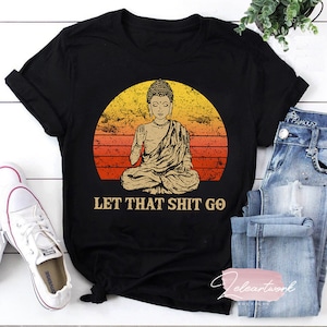 May include: Black t-shirt with a vintage-style illustration of a meditating Buddha in front of a sunset. The text "Let That Shit Go" is printed below the image.