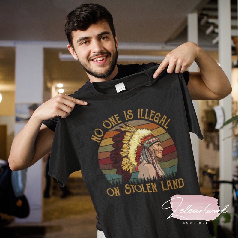 Peut inclure: Un t-shirt noir avec un graphique vintage d'un Am&eacute;rindien portant une coiffe. Le graphique est sur un fond de coucher de soleil avec le texte "No one is illegal on stolen land".