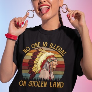 Peut inclure: T-shirt noir avec un graphique vintage d'un Am&eacute;rindien portant une coiffe. Le graphique est sur fond de coucher de soleil avec le texte "No one is illegal on stolen land".