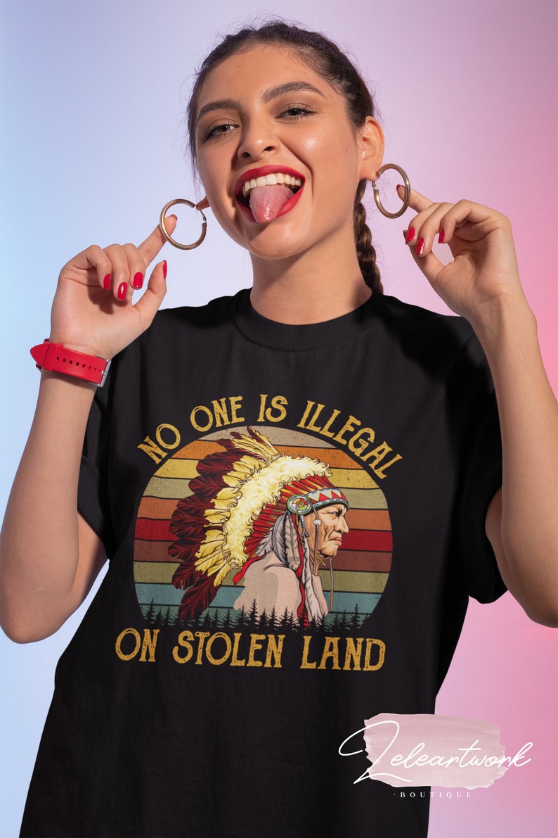 Peut inclure: T-shirt noir avec un graphique vintage d'un Am&eacute;rindien portant une coiffe. Le graphique est sur fond de coucher de soleil avec le texte "No one is illegal on stolen land".