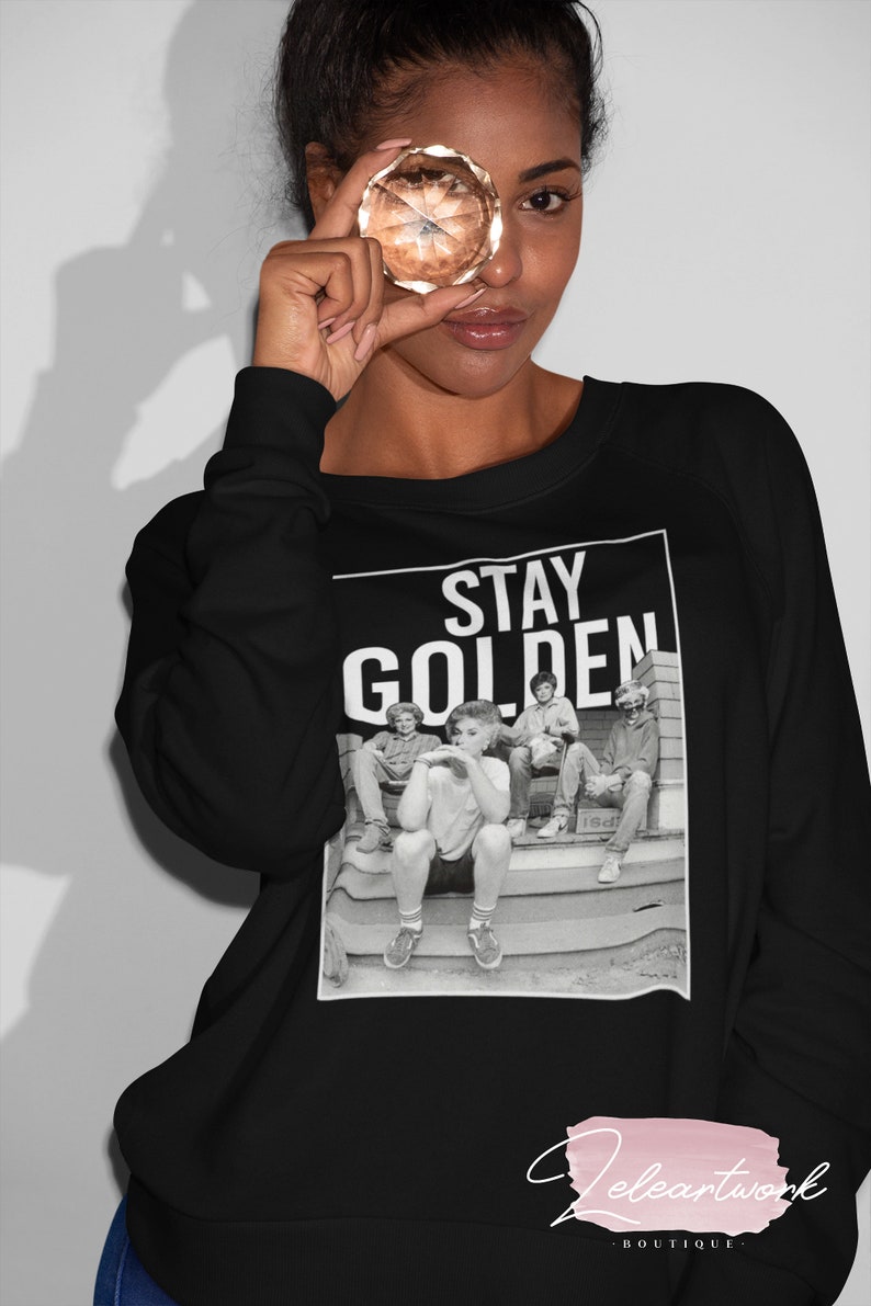 Puede incluir: Sudadera negra con un estampado gr&aacute;fico blanco de un grupo de personas sentadas en unos escalones. El gr&aacute;fico dice "Stay Golden".