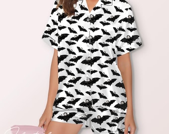 May include: White pajama set with a black bat pattern. The short-sleeved top and shorts feature a repeating design of bats with a face. The pajamas have black trim and buttons.