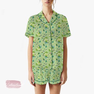 Può includere: Pigiama verde con un motivo stravagante di elementi simili a cartoni animati, tra cui foglie e baffi. Il set presenta una camicia con bottoni e colletto e pantaloncini abbinati, il tutto rifinito con bordini verdi.