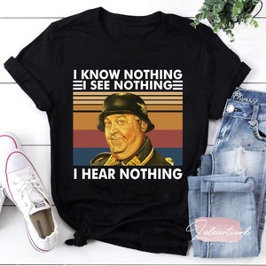 May include: Black t-shirt featuring a vintage-style graphic of a man in a military helmet with the text "I KNOW NOTHING, I SEE NOTHING, I HEAR NOTHING." The design has a retro color scheme.