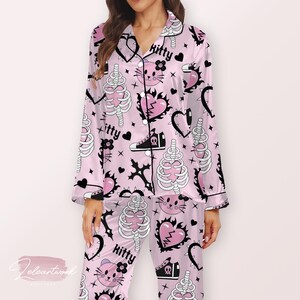 Può includere: Pigiama rosa chiaro con un motivo ripetuto di figure a forma di scheletro, cuori e la parola "kitty". Il set comprende una camicia con bottoni e pantaloni, entrambi rifiniti con bordini neri.