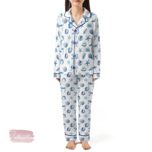 May include: White pyjama set featuring a blue seashell and starfish design. The long-sleeved top has a button-down front and navy blue trim. The matching trousers have a relaxed fit. The pyjamas are made of a soft, lightweight fabric.