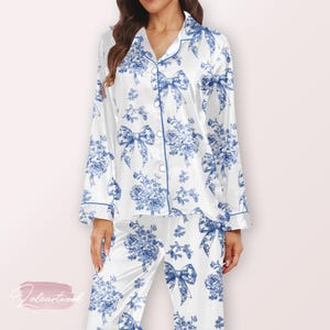 Könnte beinhalten: Weißes Satin-Pyjama-Set mit blauem Blumen- und Schleifenmuster. Das langärmlige Oberteil hat eine Knopfleiste und blaue Paspeln. Die passende Hose vervollständigt das Set und schafft ein koordiniertes Nachtwäsche-Ensemble.