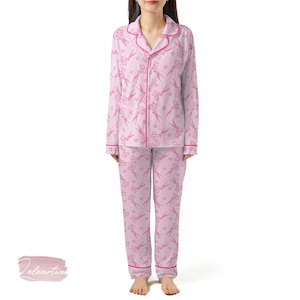 Pode incluir: Conjunto de pijama rosa claro com um padrão repetido de tesouras e corações. A parte superior de manga comprida e as calças são adornadas com um debrum rosa mais escuro. O pijama tem uma parte superior abotoada com gola.