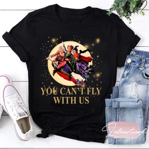 Pode incluir: Camiseta preta com um gráfico de três bruxas voando em uma vassoura na frente de uma lua cheia. O texto "You Can't Fly With Us" é impresso em dourado abaixo da imagem.