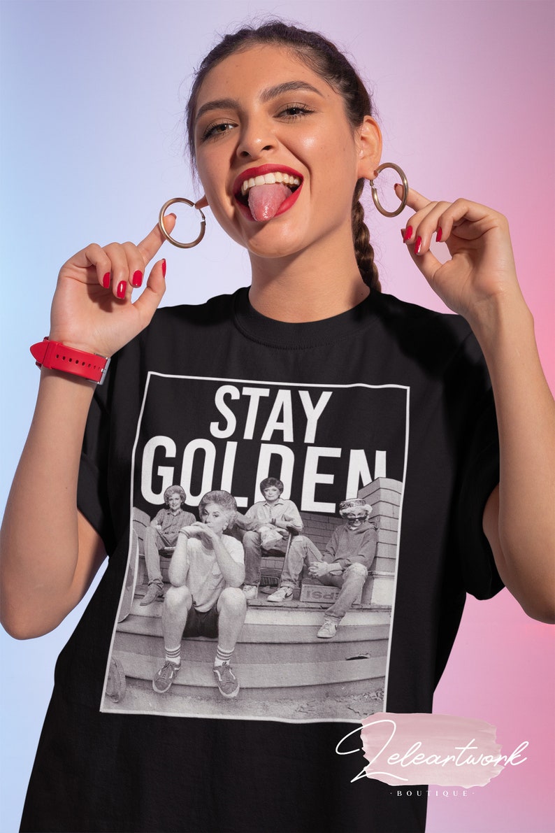 Puede incluir: Camiseta negra con un estampado gr&aacute;fico de las Golden Girls sentadas en un porche. El texto "STAY GOLDEN" est&aacute; impreso en blanco sobre la imagen.