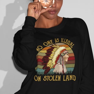 Peut inclure: Sweat-shirt noir avec un graphique vintage d'un Am&eacute;rindien portant une coiffe. Le graphique comprend un coucher de soleil et le texte "No one is illegal on stolen land."
