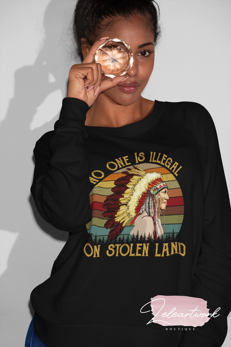 Peut inclure: Sweat-shirt noir avec un graphique vintage d'un Am&eacute;rindien portant une coiffe. Le graphique comprend un coucher de soleil et le texte "No one is illegal on stolen land."