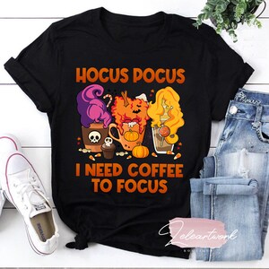 Pode incluir: Camiseta preta com um design temático de Halloween com três bruxas segurando canecas de café. O texto "Hocus Pocus I Need Coffee To Focus" está impresso na camiseta.