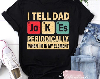Op de afbeelding: Zwart T-shirt met een vintage-stijl grafische afbeelding met de tekst "Ik vertel mijn vader regelmatig grappen wanneer ik in mijn element ben". De afbeelding bevat de chemische symbolen voor "Jo", "K" en "Es" in kleurrijke vakjes.