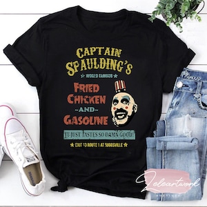Puede incluir: Camiseta negra con un gráfico de estilo vintage de Captain Spaulding's Fried Chicken and Gasoline. El gráfico presenta una caricatura de Captain Spaulding con el texto "World Famous" y "It just tastes so damn good! Exit 13 Route 1 at Suggsville."