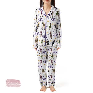 May include: White pajama set with a repeating pattern of stylized figures, crowns, and the word "SIX" in purple. The set includes a button-down top with purple trim and matching pants. The pajamas are made of a soft, comfortable material.