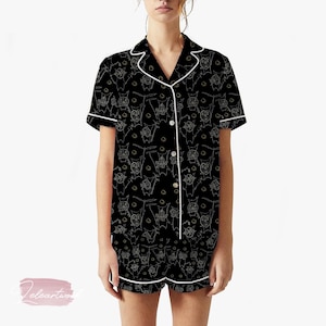 Puede incluir: Conjunto de pijama negro con un patrón de contorno blanco de personajes de dibujos animados. La parte superior de manga corta y los pantalones cortos tienen ribetes y botones blancos. El pijama está hecho de un material suave y cómodo.