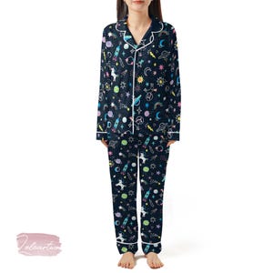 May include: Navy blue pyjama set with a colourful space-themed pattern. The long-sleeved top and trousers feature rockets, stars, planets, and unicorns. White piping trims the collar, cuffs, and pockets. The pyjamas are designed for comfort and style.