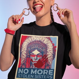 May include: Black t-shirt with a graphic of a Native American woman in a headdress, red face paint, and a red handprint. The text "NO MORE STOLEN SISTERS" is below the image. The woman in the image is wearing gold hoop earrings.