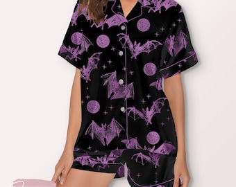 May include: Black pajama set with a purple bat and moon pattern. The short-sleeved top has a notched collar and button closure. The shorts have an elastic waistband. The set is trimmed with white piping.