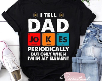 Op de afbeelding: Zwart T-shirt met een wit grafisch ontwerp met de tekst "I tell Dad Jokes Periodically but only when I'm in my element". Het grafische ontwerp bevat een gestileerd periodiek systeem met de gemarkeerde elementen "Jo", "K" en "Es".