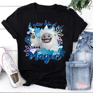 Puede incluir: Camiseta negra con una criatura blanca similar a un yeti con ojos azules y boca azul. La criatura está rodeada de copos de nieve y el texto "A new kind of magic".