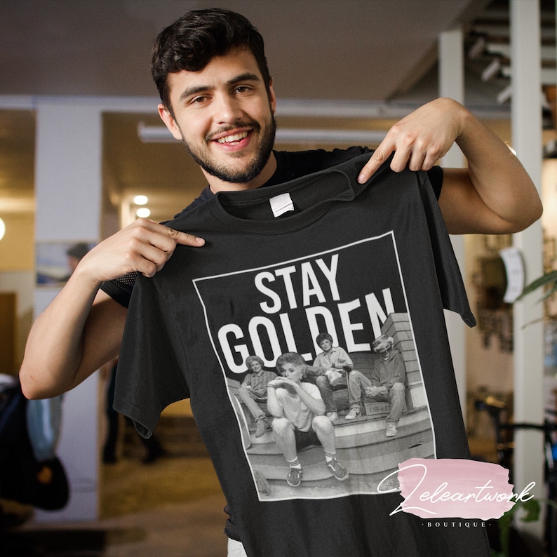Puede incluir: Camiseta negra con un estampado gr&aacute;fico blanco de un grupo de personas sentadas en unos escalones. El gr&aacute;fico incluye el texto "Stay Golden".