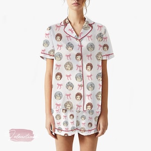 Könnte beinhalten: Weißes Pyjama-Set mit einem sich wiederholenden Muster aus illustrierten Gesichtern und rosa Schleifen. Das kurzärmelige Oberteil und die Shorts haben einen rosa Besatz. Das Pyjama-Set ist aus einem weichen, bequemen Material.