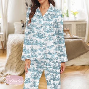 Pode incluir: Conjunto de pijama toile de Jouy branco e azul. A parte superior de manga comprida tem uma frente abotoada e uma gola entalhada. As calças são compridas e têm uma estampa combinando. O conjunto apresenta um design de paisagem azul sobre fundo branco.