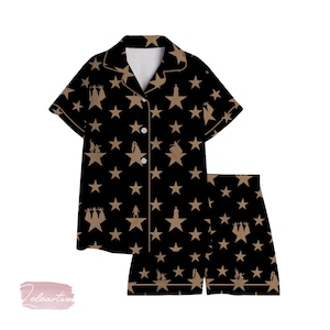 Puede incluir: Conjunto de pijama negro con un estampado de estrellas y siluetas. La parte superior de manga corta tiene cuello y cierre de botones. Los pantalones cortos tienen cintura elástica. El diseño presenta estrellas y siluetas doradas.