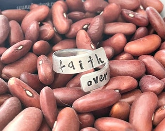 May include: A silver ring with the words "faith over" wrapped around the band. The ring is sitting on a bed of red kidney beans.