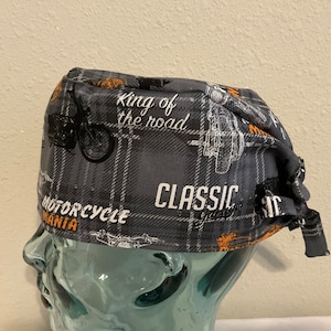 May include: Gray and orange patterned surgical scrub hat with a motorcycle theme. The hat features the text "King of the Road", "Motorcycle Mania", and "Classic Garage".