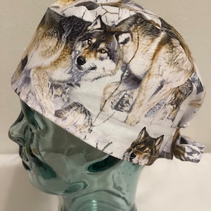 May include: A white and gray patterned cap featuring a wolf design. The cap is made of fabric and has a tie closure at the back. The wolf design includes shades of brown, gray, and black.